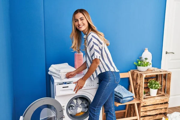 在洗衣房 年轻的金发女人满脸笑容 自信地把洗涤剂倒在洗衣机上 — 图库照片