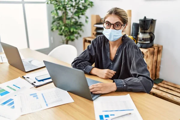 Young blonde woman working at the office wearing safety mask scared and amazed with open mouth for surprise, disbelief face