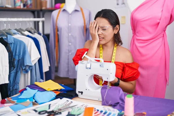 Hispanic young woman dressmaker designer using sewing machine yawning tired covering half face, eye and mouth with hand. face hurts in pain.