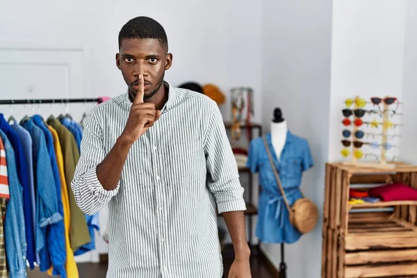 Young african american man working as manager at retail boutique asking to be quiet with finger on lips. silence and secret concept.