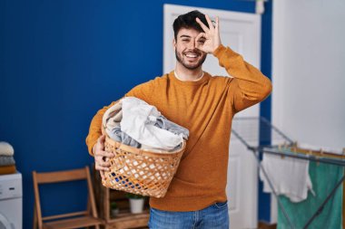 Çamaşır odasında sakallı, çamaşır sepetini tutan İspanyol bir adam gülümsüyor. 