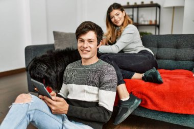 Evde dokunmatik ped kullanan, köpekle mutlu gülümseyen beyaz bir çift..