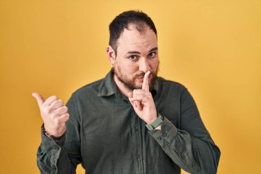 Plus size hispanic man with beard standing over yellow background asking to be quiet with finger on lips pointing with hand to the side. silence and secret concept. 