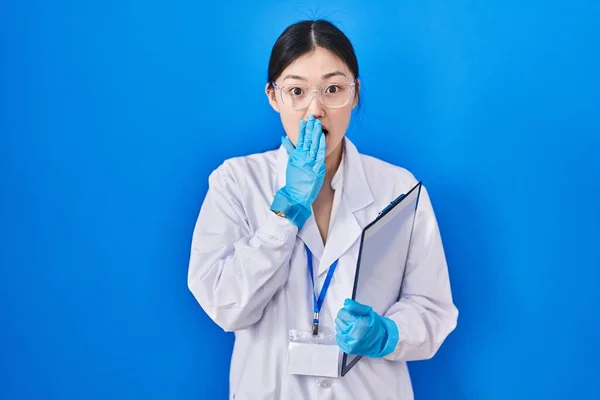 Chinese young woman working at scientist laboratory afraid and shocked, surprise and amazed expression with hands on face