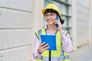 Young hispanic woman architect talking on the smartphone using touchpad at street