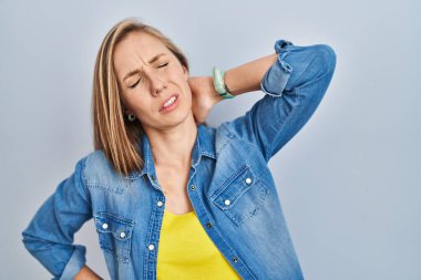 Mavi arka planda duran genç sarışın kadın boyun ağrısı çekiyor, eliyle boynuna dokunuyor, kas ağrısı çekiyor. 