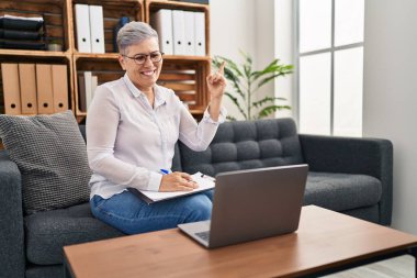 Orta yaşlı bir kadın danışma ofisinde online seans yapıyor. Gülümsüyor, mutlu bir şekilde işaret ediyor. 