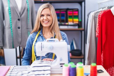 Blonde woman dressmaker designer using sew machine winking looking at the camera with sexy expression, cheerful and happy face. 