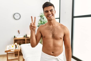 İspanyol asıllı genç adam spa merkezinde üstsüz duruyor. İki numarayı gösterip işaret ediyor. Kendinden emin ve mutlu bir şekilde gülümsüyor.. 