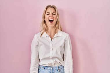 Young caucasian woman wearing casual white shirt over pink background angry and mad screaming frustrated and furious, shouting with anger. rage and aggressive concept. 