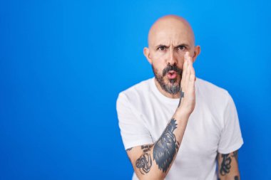 Mavi arka planda dikilen İspanyol dövmeli adam ağzının üstünde gizli dedikodular söylüyor, kötü niyetli konuşmalar fısıldıyor. 