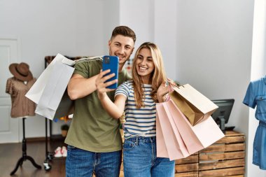 Young hispanic customer couple holding shopping bags and make selfie by the smartphone at clothing store.