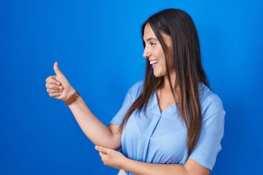 Young brunette woman standing over blue background looking proud, smiling doing thumbs up gesture to the side 