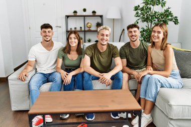 Mutlu bir şekilde gülümseyen bir grup genç arkadaş evdeki kanepede oturuyor..