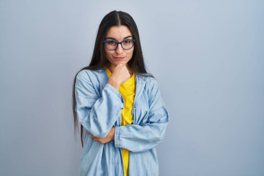 Mavi arka planda duran genç İspanyol kadın kameraya kendinden emin bir şekilde bakıyor kollarını kavuşturmuş ve çenesini kaldırmış gülümsüyor. Olumlu düşünmek. 