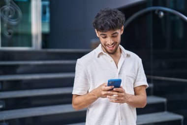 Genç Arap adam sokakta akıllı telefon kullanarak kendinden emin gülümsüyor.