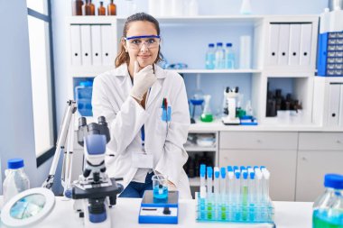 Young hispanic woman working at scientist laboratory looking confident at the camera with smile with crossed arms and hand raised on chin. thinking positive. 