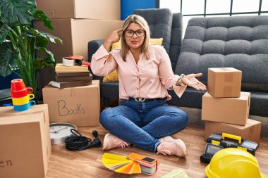 Young hispanic woman moving to a new home sitting on the floor confused and annoyed with open palm showing copy space and pointing finger to forehead. think about it. 