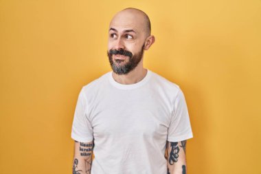 Sarı arka planda dikilmiş, gülümseyen ve gözlerini kaçıran İspanyol dövmeli genç adam.. 