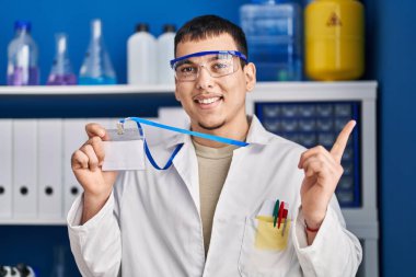 Young arab man working at scientist laboratory holding id card smiling happy pointing with hand and finger to the side 