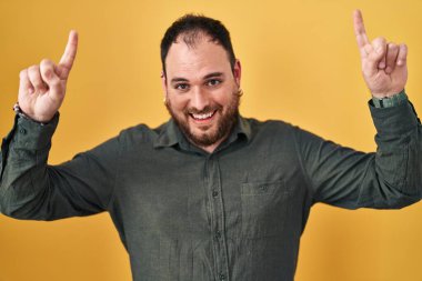 Plus size hispanic man with beard standing over yellow background smiling amazed and surprised and pointing up with fingers and raised arms. 