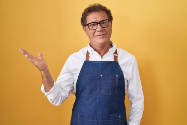 Middle age hispanic man wearing professional cook apron smiling cheerful presenting and pointing with palm of hand looking at the camera. 