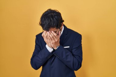 Handsome latin man standing over yellow background with sad expression covering face with hands while crying. depression concept. 