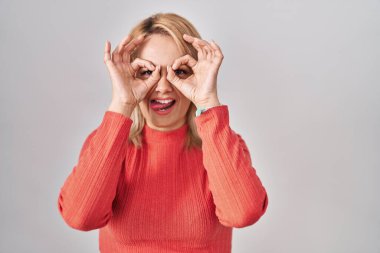 Sarışın kadın izole edilmiş arka planda dikiliyor dürbün gibi hareket ediyor, gözleri parmaklarından bakıyor. çılgın ifade. 