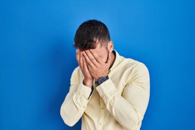 Handsome hispanic man standing over blue background with sad expression covering face with hands while crying. depression concept. 