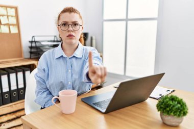 Ofiste çalışan genç kızıl saçlı kadın bilgisayarı kullanarak parmağını kaldırmış ve kızgın bir ifadeyle işaret ediyor. 