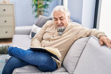 Orta yaşlı, kır saçlı, kitap okuyan adam evdeki kanepede oturuyor.