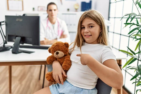 Blonde little girl at pediatrician clinic with female doctor smiling happy pointing with hand and finger