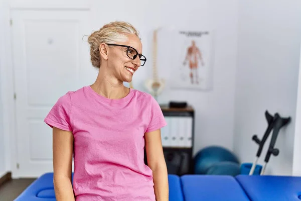 Middle age blonde woman at pain recovery clinic looking away to side with smile on face, natural expression. laughing confident.