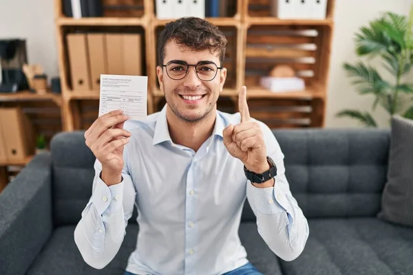 Young hispanic man holding covid record card smiling with an idea or question pointing finger with happy face, number one