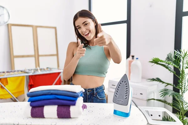 洗濯室で若いヒスパニック系の女性のアイロン服をカメラに幸せと面白い顔を指を指している 良いエネルギーと振動は — ストック写真
