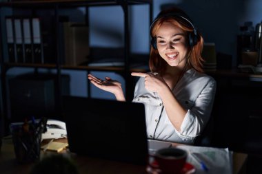 Geceleri ofiste çalışan beyaz bir kadın hayret içinde kameraya bakıp gülümserken eliyle ve parmağıyla işaret ediyor.. 