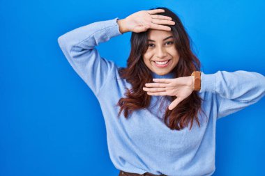 Hispanic young woman standing over blue background smiling cheerful playing peek a boo with hands showing face. surprised and exited 