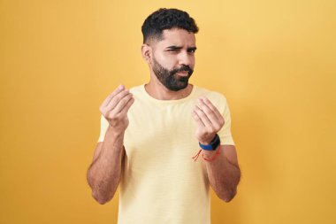 Hispanic man with beard standing over yellow background doing money gesture with hands, asking for salary payment, millionaire business 