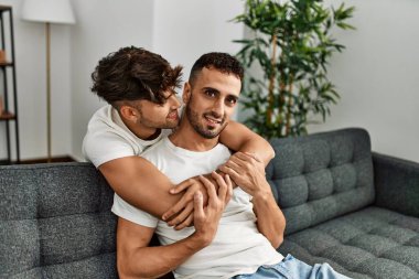 İki İspanyol erkek, kendilerine güvenerek gülümsüyor ve evde birbirlerine sarılıyorlar.