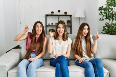 Group of three hispanic girls sitting on the sofa at home pointing finger up with successful idea. exited and happy. number one. 