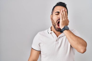 Sakallı, beyaz arka plan üzerine günlük kıyafetler giyen, yarı yüz, göz ve ağız kaplamasından yorgun düşmüş genç İspanyol bir adam. Yüzüm acıyor.. 