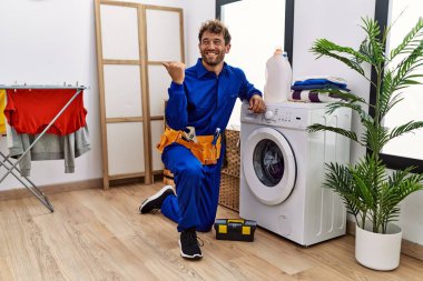 Çamaşır makinesinde çalışan genç İspanyol bir adam baş parmağını yukarı kaldırıyor ve ağzı açık gülümsüyor. 