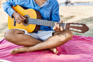 Klasik gitar çalan bir adam kumsalda oturuyor..