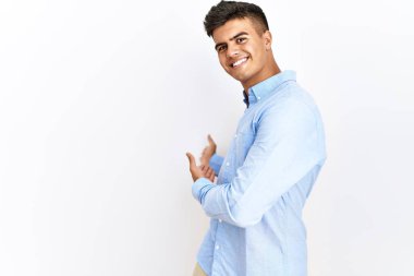 İş gömleği giyen genç İspanyol adam izole edilmiş arka planda duruyor ve doğal bir şekilde gülümseyerek içeri girmeye davet ediyor. 