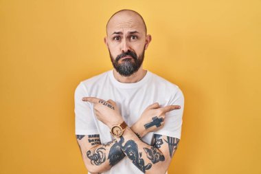 Sarı arka planda dikilen İspanyol dövmeli genç adam her iki tarafı da parmaklarıyla işaret ediyor, farklı yönlere. 