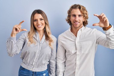 Young couple standing over blue background smiling and confident gesturing with hand doing small size sign with fingers looking and the camera. measure concept. 