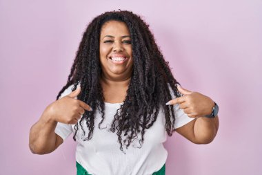 Plus size hispanic woman standing over pink background looking confident with smile on face, pointing oneself with fingers proud and happy. 