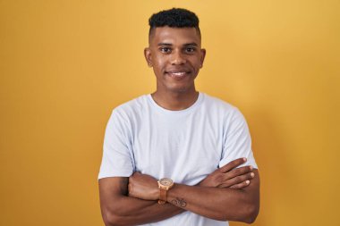 Sarı arka planda duran İspanyol genç adam kollarını kavuşturup kameraya bakarken gülümsüyor. pozitif kişi. 