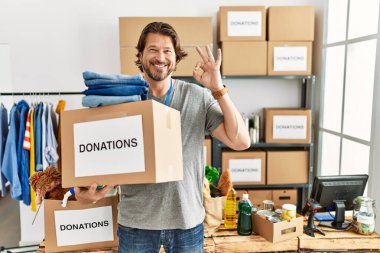 Gönüllü olarak hayır işi için bağış kutusunu tutan yakışıklı orta yaşlı adam gülümseyerek el ve parmaklarıyla tamam işareti yapıyor. başarılı ifade. 