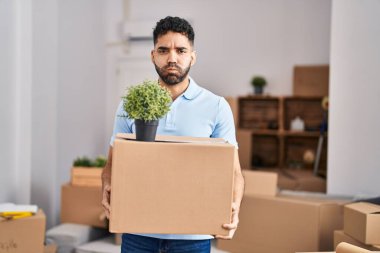 Hispanic man with beard moving to a new home holding box puffing cheeks with funny face. mouth inflated with air, catching air. 
